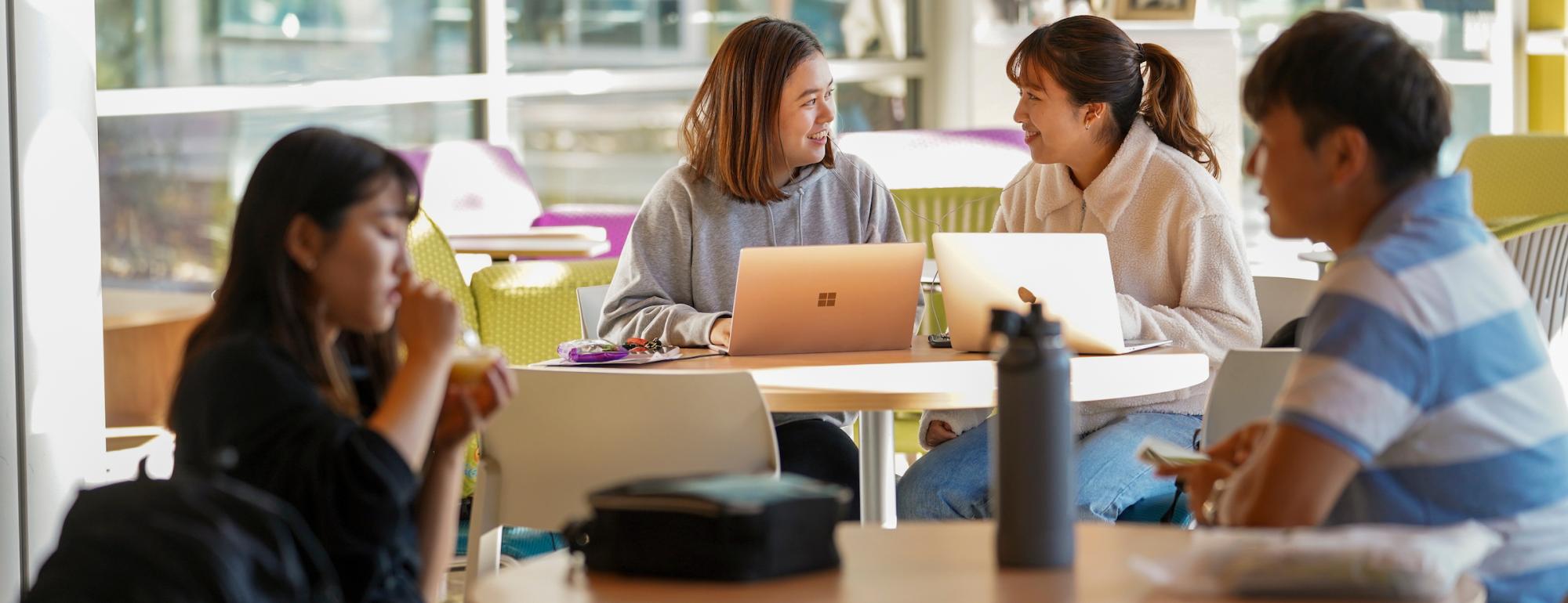 A picture of 4 students who are studying and talking at a table. Two are out of focus on the outer edge and the center two are in focus.